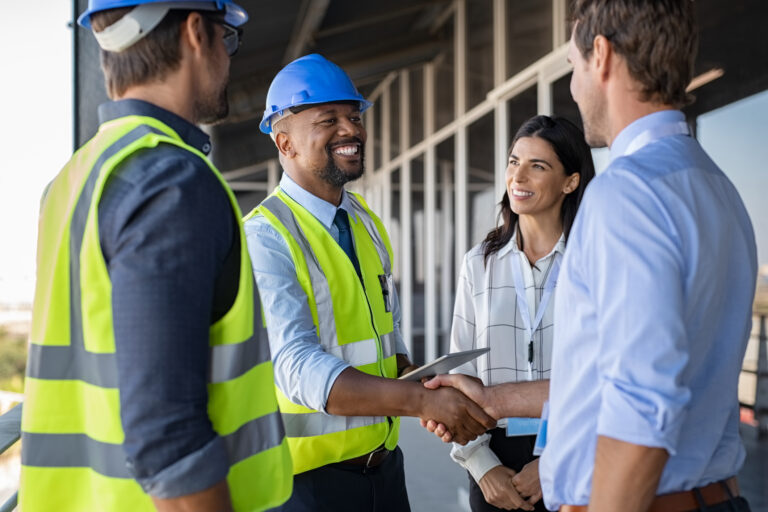 Four construction workers. The two on the left are wearing helmets and yellow vests. The two at the right are wearing button up shirts. The middle two are shaking hands as thought they made an agreement.
